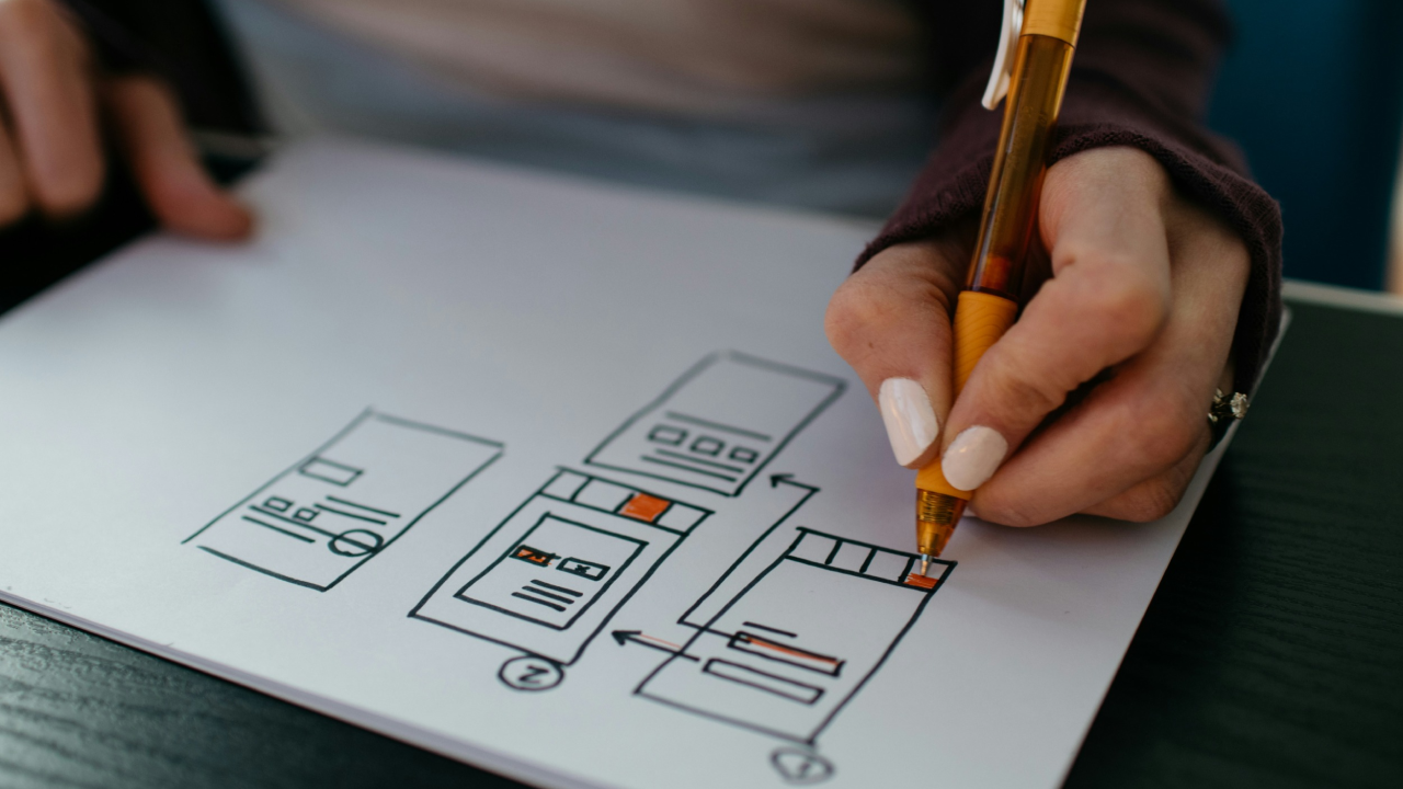 Close-up of a person's hand drawing a website wireframe with boxes and arrows on paper.