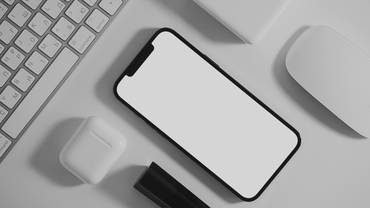 Flat lay workspace with blank iPhone mockup, keyboard, AirPods and mouse on white surface.