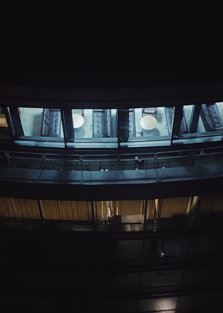 View through lit-up windows in a high-rise building showing empty tables and chairs at night.