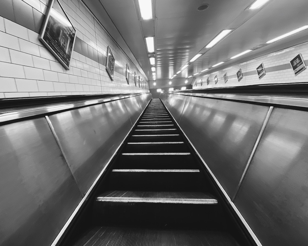 Black and white photo of metro station escalator leading upwards with bright lighting.